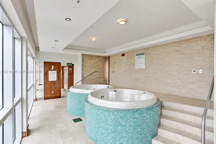 Spa facility featuring dual circular jetted tubs with turquoise tile surrounds, natural stone wall cladding, recessed lighting, and tiered stone steps with a metal handrail