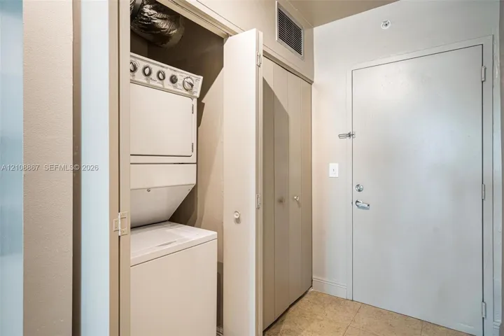 Laundry closet featuring a stacked washer and dryer, neutral wall paint, tile flooring, and a solid-core entry door with a metal deadbolt