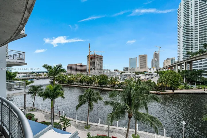 Private balcony with metal railings overlooking a waterfront promenade, mature palm trees, and an urban cityscape