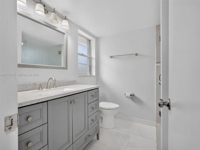 Bathroom featuring a grey shaker-style vanity, stone-composite countertop with an integrated basin, multi-drawer storage, polished nickel-finish hardware, and large-format porcelain floor tile