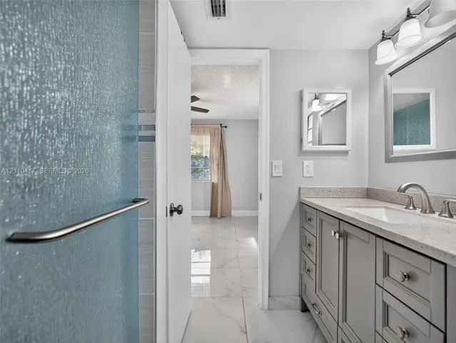 Bathroom featuring a light-toned vanity with stone-surface countertop, rectangular undermount sink, three-light wall fixture, and marble-finish flooring
