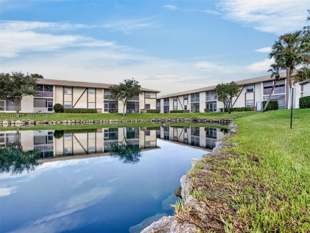 Waterfront multi-story residential building with stone-lined shore, manicured lawn, and mature palm trees