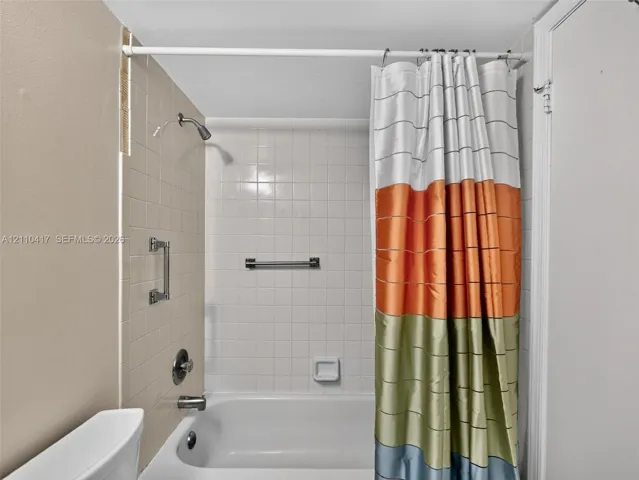 Bathtub and shower combination featuring a porcelain finish, wall-mounted shower head, integrated grab bar, and neutral square wall tiling