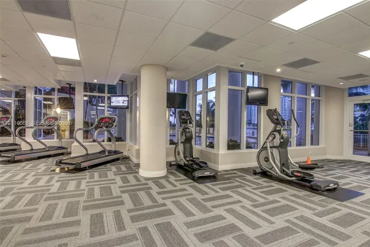 Fitness center featuring expansive floor-to-ceiling windows, high-contrast patterned commercial carpeting, recessed lighting, and structural support columns