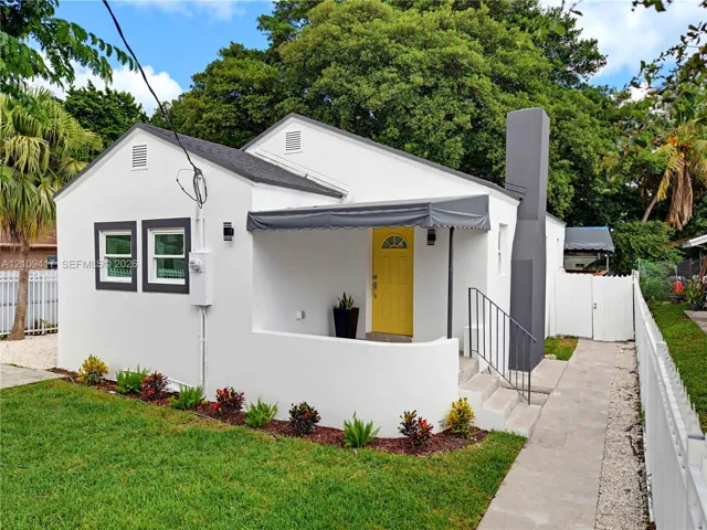 Exterior featuring a stucco facade, covered front entry with a metal awning, prominent exterior chimney, and a white perimeter fence