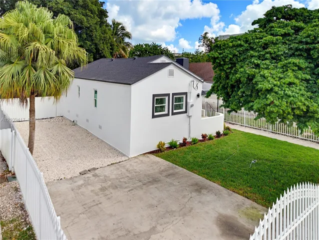 Exterior featuring a white stucco facade, dark shingle roof, concrete driveway, gravel side yard, and enclosed lawn area with perimeter fencing