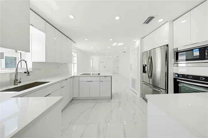 Contemporary kitchen featuring high-gloss white cabinetry, white stone-finish countertops, stainless steel appliances, and large-format porcelain tile flooring with grey veining