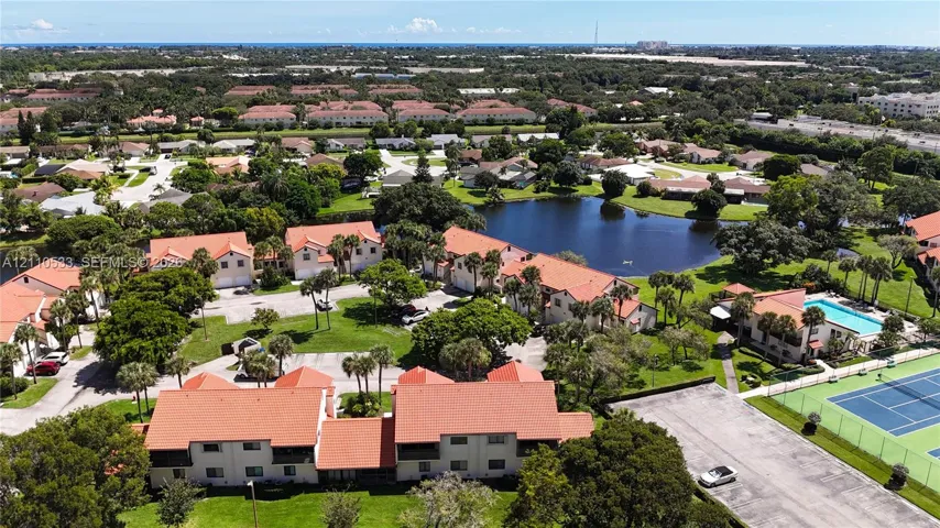 Aerial perspective of the residential complex featuring tile roofing, a central pond, a swimming pool, and a hard-court tennis facility set within landscaped grounds
