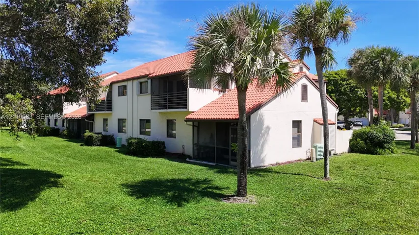 Two-story multi-family exterior featuring light-toned stucco siding, a tiled roof, screened balconies, and mature palm landscaping