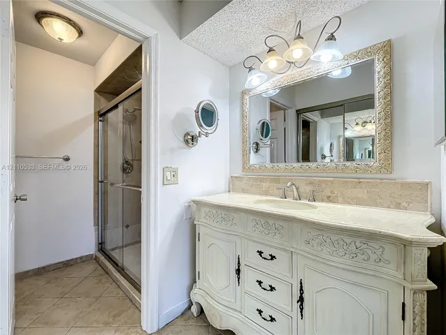 Vanity featuring ornate cabinetry, natural stone countertop, and integrated sink with widespread faucet, accompanied by a shower enclosure with tile surround and a wall-mounted magnifying mirror