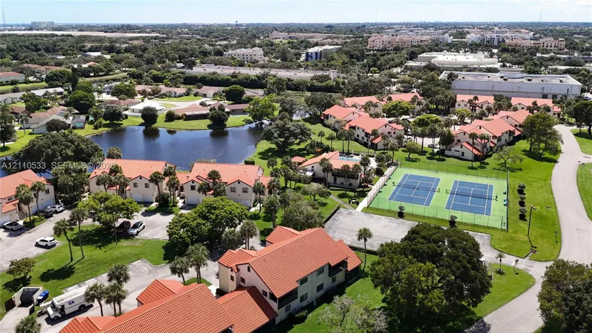 Aerial perspective of the community featuring dual tennis courts, a central pond, multi-story buildings with terracotta-style roof tiles, and mature landscaping