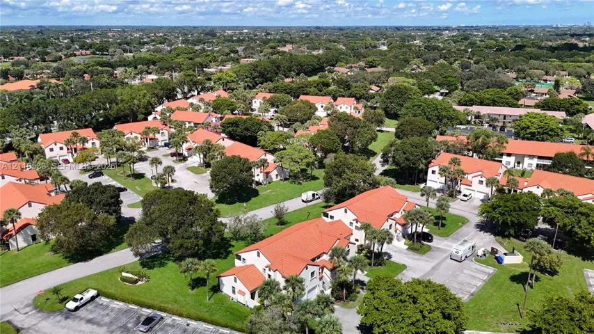 Aerial perspective of a multi-unit residential community featuring clay tile roof systems, stucco exteriors, mature trees, and paved parking surfaces