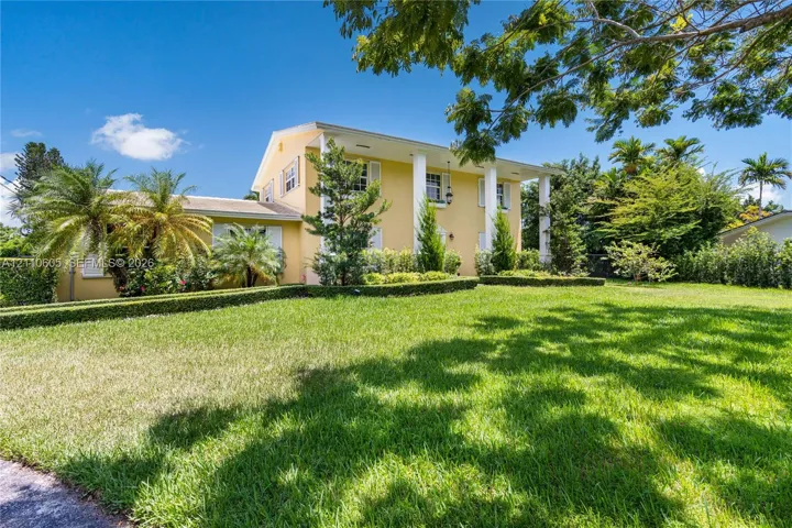 Two-story exterior featuring a columned front portico, light-toned stucco siding, and meticulously manicured hedging on an expansive lawn with mature palm trees