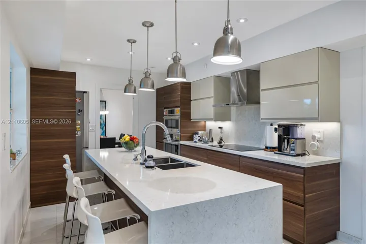Kitchen featuring a waterfall-edge center island, wood-finish cabinetry, light-toned solid surface countertops, stainless steel pendant lighting, and a stainless steel wall-mounted range hood