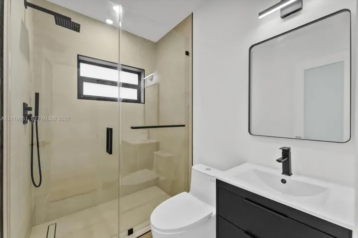 Bathroom featuring a glass-enclosed shower with a rainfall showerhead, a black vanity with a white countertop and rectangular sink, a black-framed mirror, and matte black fixtures