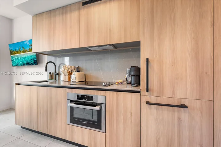 Modern cabinetry with a light wood-finish, sleek stone countertops, integrated stainless-steel wall oven, matte black hardware, and an undermount sink with a matching matte black faucet