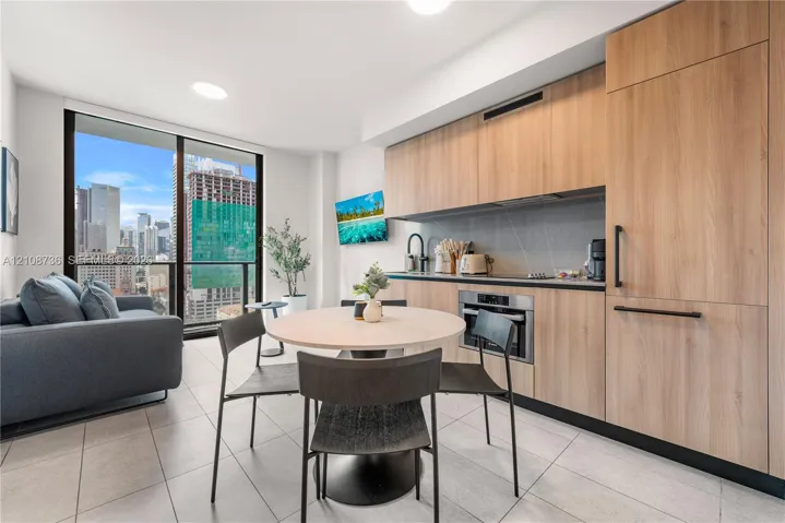 Contemporary kitchen featuring light wood-finish cabinetry, integrated panel-ready appliances, stone-composite backsplash, floor-to-ceiling glass sliding door, and neutral-toned tile flooring