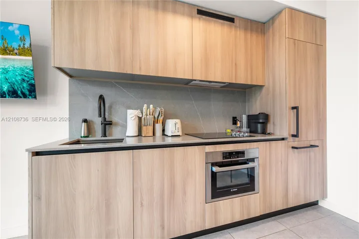Kitchen featuring flat-panel wood-finish cabinetry, stone-composite countertop, full-height stone backsplash, undermount sink with matte black faucet, and built-in stainless steel appliances