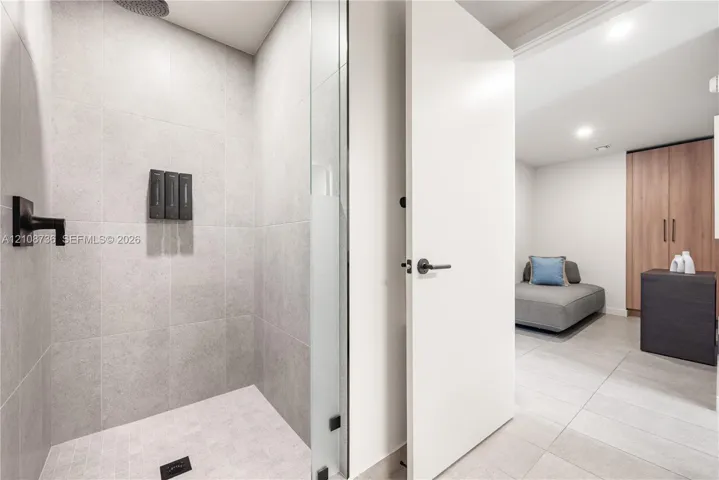 Walk-in shower featuring matte black fixtures, rainfall showerhead, and floor-to-ceiling large-format tile, complemented by a glass panel enclosure and light-toned tile flooring