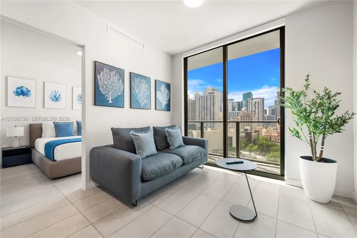Floor-to-ceiling sliding glass doors, neutral tile flooring, and a private balcony featuring urban scenery