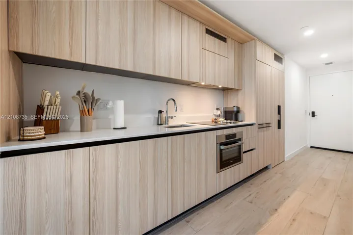 Modern kitchen featuring light wood-finish cabinetry, solid surface countertops, built-in stainless steel appliances, and wide-plank wood-finish flooring