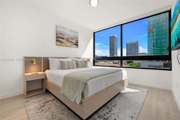 Floor-to-ceiling windows featuring urban scenery, built-in wooden bed frame with integrated headboard, and light wood-finish flooring
