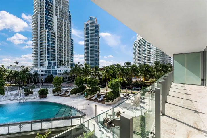 Covered balcony featuring stone flooring and a glass panel railing overlooking a swimming pool with paved decking and mature palm trees