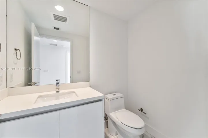 White vanity with solid-surface countertop, rectangular integrated sink, chrome single-handle faucet, full-width mirror, and dual-flush toilet