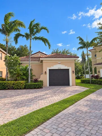 Exterior facade featuring a tile roof, stucco finish, single-car garage, and a paved stone driveway complemented by established palm trees and manicured hedges