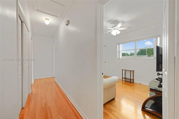 Interior hallway featuring wood-finish flooring, two-panel interior doors, and a ceiling-mounted light fixture, opening into a room equipped with a multi-blade ceiling fan and a rectangular window