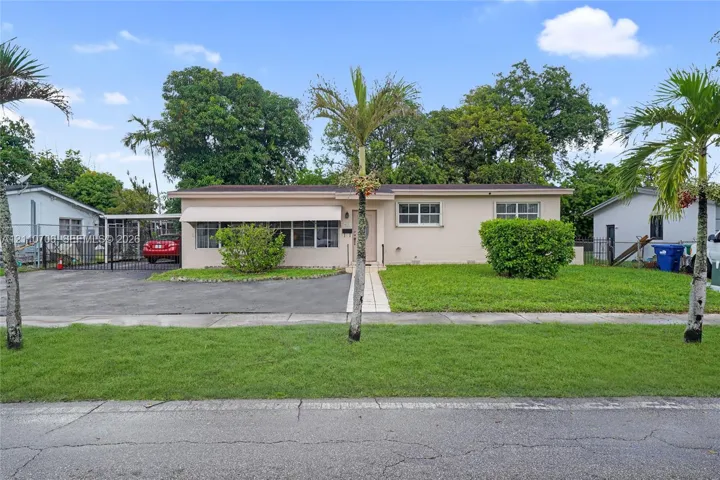 Single-story exterior featuring a light-toned masonry facade, asphalt driveway, covered metal carport, and manicured landscaping