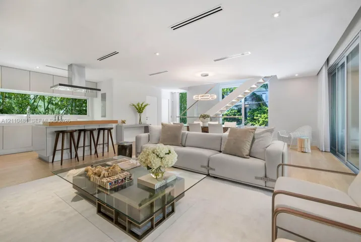 Open-concept interior featuring light wood-finish flooring, linear recessed lighting, a modern floating staircase with glass railings, large horizontal windows, and a stainless steel kitchen ventilation hood