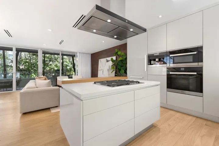 Kitchen featuring flat-panel cabinetry, center island with a gas cooktop, built-in wall ovens, stainless steel ceiling-mounted vent hood, and wood-finish flooring