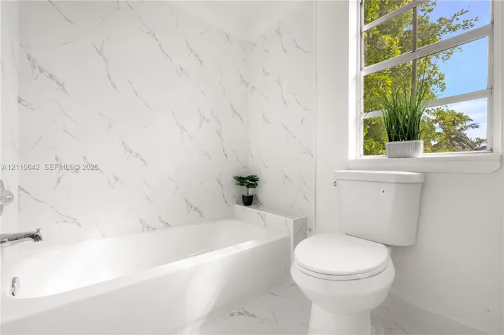 Full-height stone-patterned tile wall surround, integrated bathtub with polished chrome fixtures, white ceramic toilet, and large window with exterior greenery
