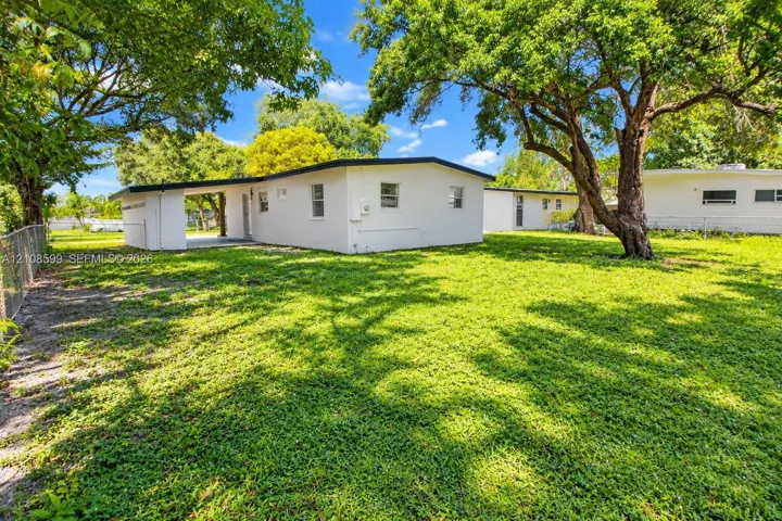 Exterior structure featuring a white stucco finish, covered side porch, mature trees, and chain-link perimeter fencing