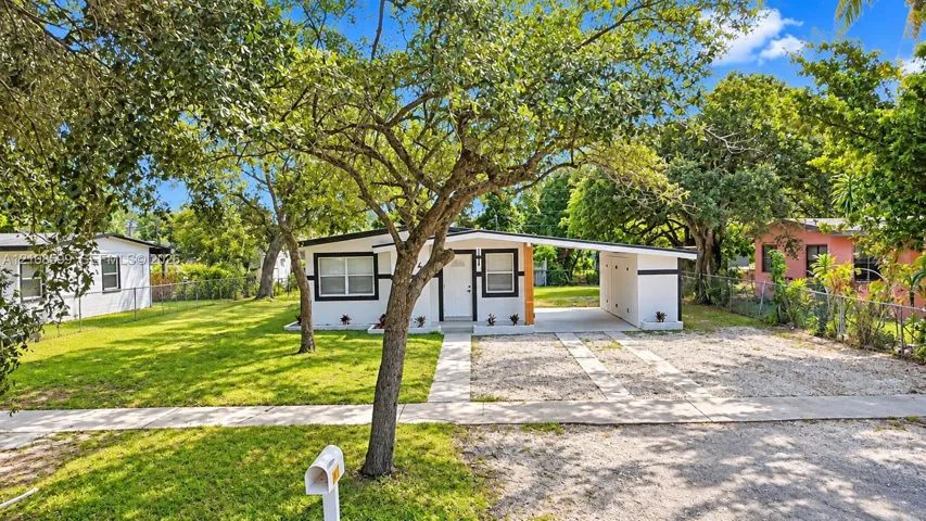 Single-story exterior featuring a flat roofline, attached carport, gravel driveway, and a large front lawn with mature trees