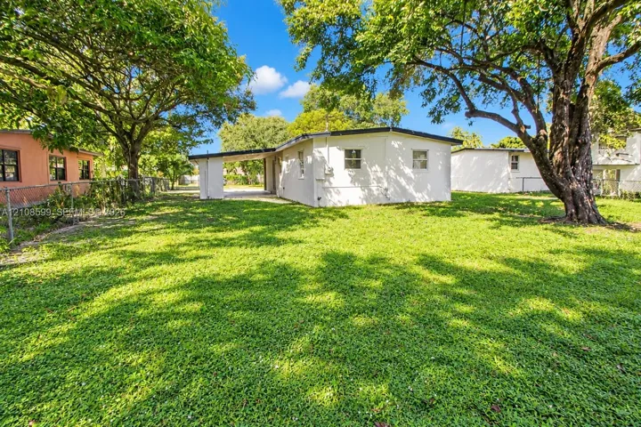 Exterior featuring a white stucco facade, covered carport, chain-link perimeter fencing, and mature trees across the grounds