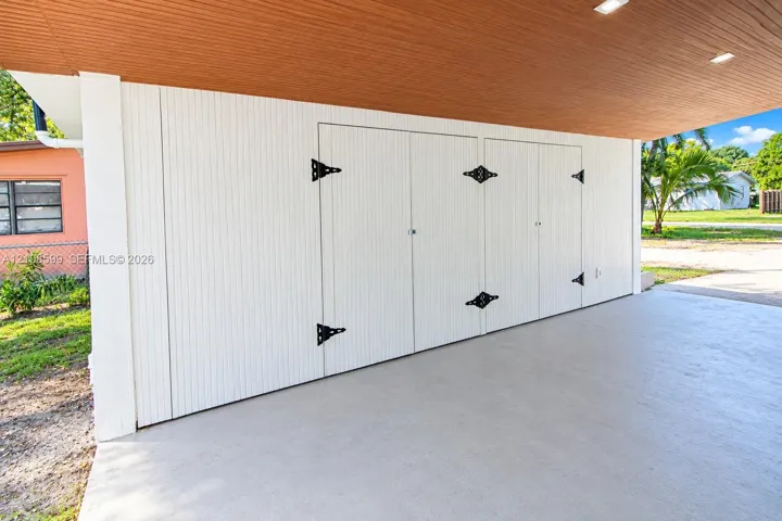 Covered carport featuring a finished wood-tone ceiling with recessed lighting, smooth concrete flooring, and white vertical-paneled storage cabinetry with decorative black iron hardware