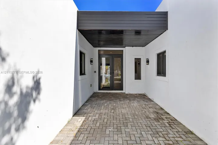 Covered entryway featuring a herringbone-patterned paver floor, minimalist smooth-stucco exterior walls, dark wood-finish ceiling paneling, glass-paneled double doors, and wall-mounted exterior sconces
