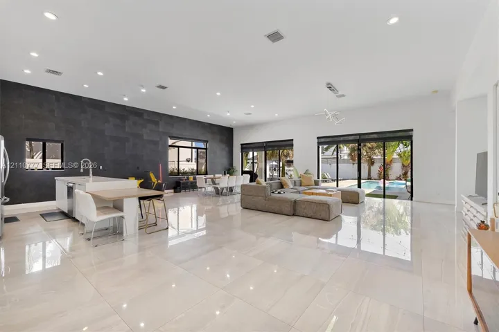Open-concept living space featuring high-gloss tile flooring, recessed lighting, a statement dark tile accent wall, an island kitchen with built-in cabinetry, and multi-panel glass sliding doors