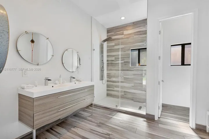 Double vanity with integrated sinks, wall-mounted faucets, wood-finish cabinetry, walk-in shower with horizontal tile surround, and wood-finish flooring