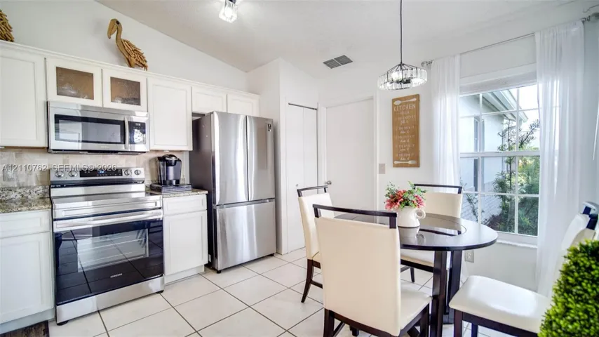 Kitchen featuring white shaker-style cabinetry, stone-finish countertops, stainless steel appliances, ceramic tile flooring, and a crystal-accented pendant light fixture