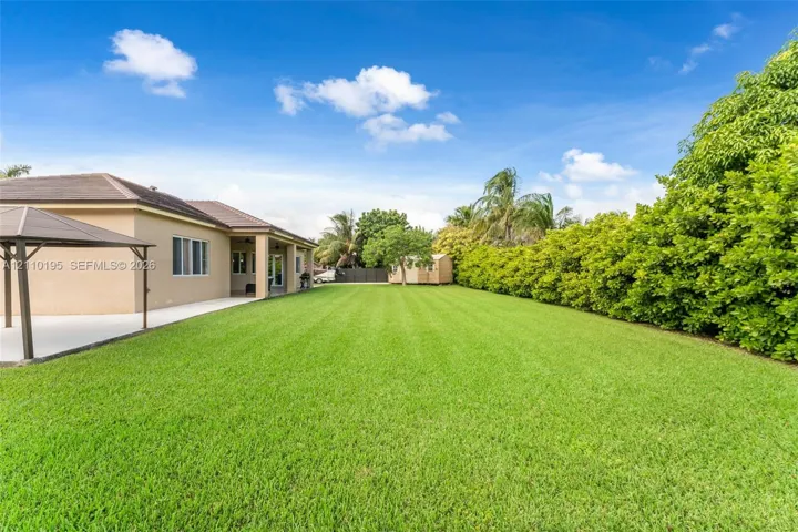 Expansive backyard featuring a level lawn, concrete patio, metal-frame gazebo, and mature perimeter hedging
