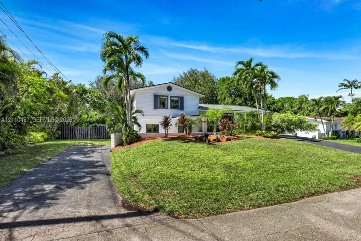 Two-story residential exterior featuring horizontal siding, dark window shutters, an asphalt driveway, and mature landscaping with palm trees