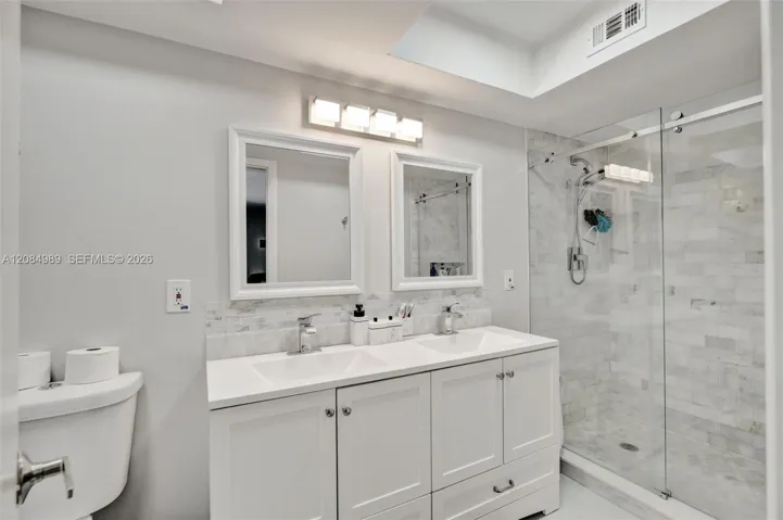 Double vanity with rectangular undermount sinks, stone-finish countertops, framed mirrors, glass-enclosed shower with tiled surround, and recessed ceiling lighting