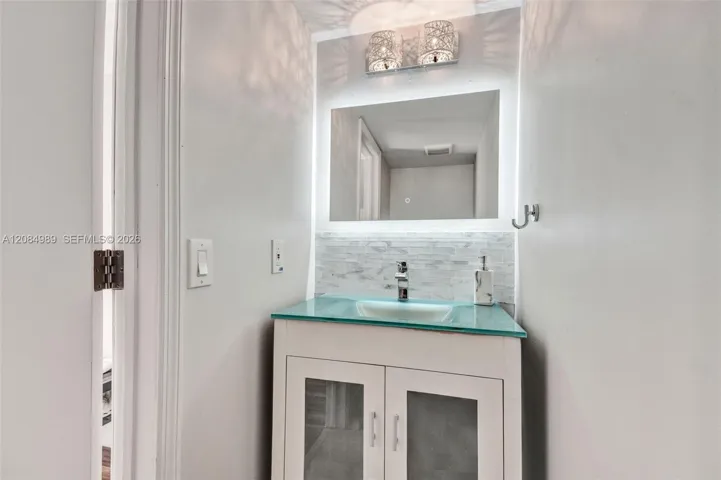 Vanity featuring a glass countertop with an integrated basin, white cabinetry with frosted glass inserts, LED-backlit mirror, and a textured tile backsplash