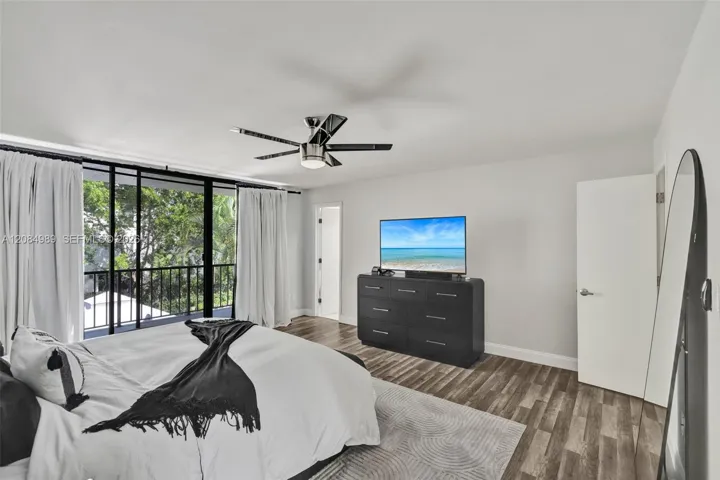 Floor-to-ceiling glass sliding doors with black framing, wood-finish flooring, metal ceiling fan with integrated lighting, and access to a railed balcony