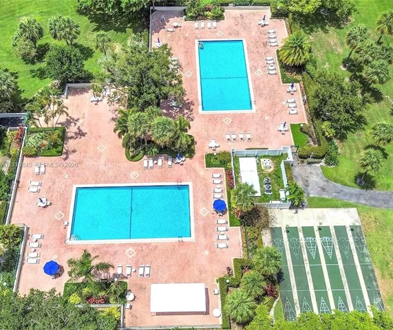 Recreational area featuring two rectangular swimming pools, expansive paved sun deck, and multiple shuffleboard courts surrounded by mature landscaping