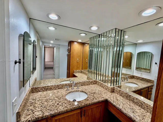 Stone-surface countertop with integrated oval basin, natural wood-finish cabinetry, floor-to-ceiling mirror paneling, and recessed ceiling lighting