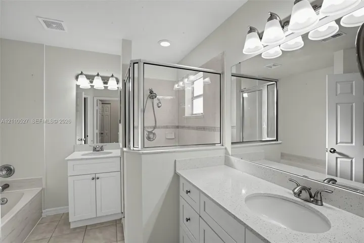 Bathroom featuring dual white shaker-style cabinetry, light-toned stone-finish countertops, an enclosed glass-paneled shower with tiled surround, and large-scale vanity mirrors with multi-bulb light fixtures